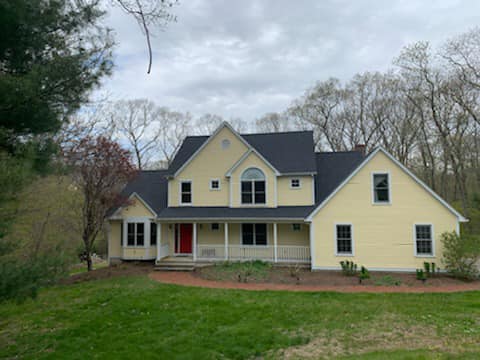 A yellow two-story house with a dark gray roof installed by a trusted roofing contractor in New London County boasts a red front door and a white porch. Nestled among trees, it is framed by grassy lawns under an overcast sky, creating a serene CT woodland setting.