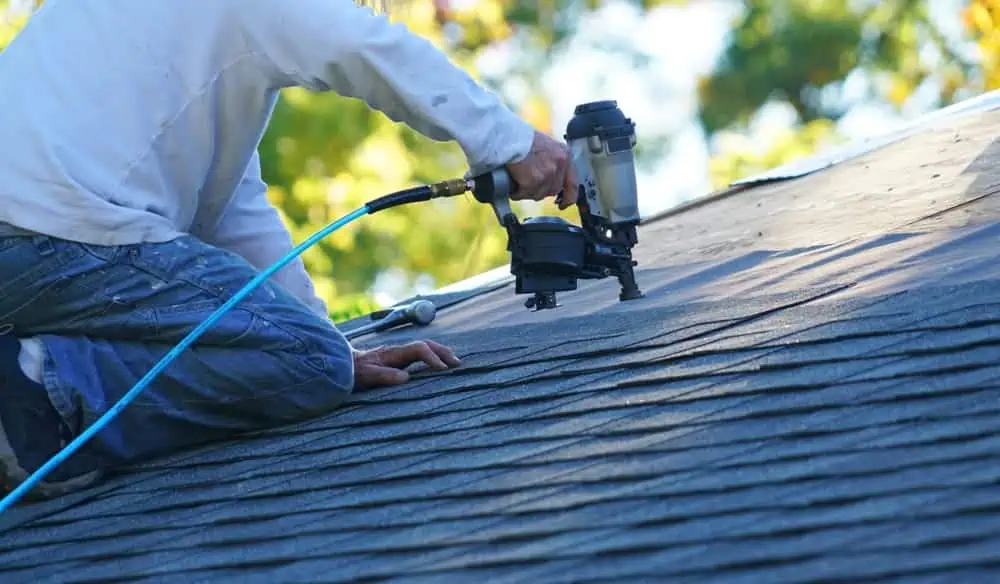 A person kneels on a roof in New London County, expertly using a nail gun for roof installation. They wear jeans and a light-colored shirt, with a blue air hose connected to the tool. The background is blurred, showcasing green and yellow foliage typical of CT landscapes.