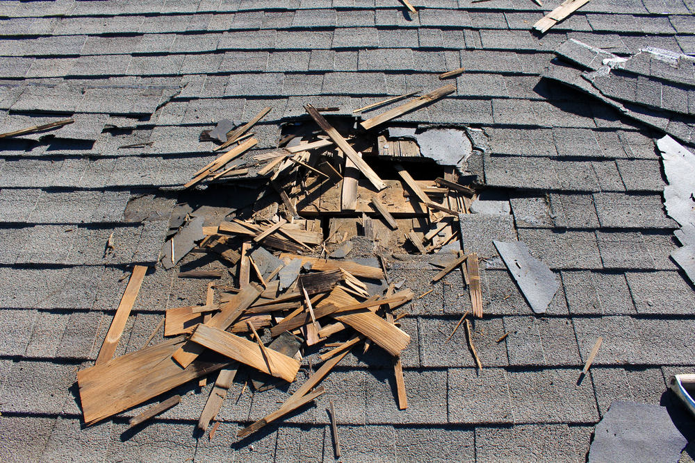 A damaged roof with gray shingles has a large hole. Wooden debris is scattered around the opening, revealing underlying materials. Other shingles are cracked and misaligned, indicating severe damage. A roofing contractor in New London County, CT could expertly handle such extensive repairs.