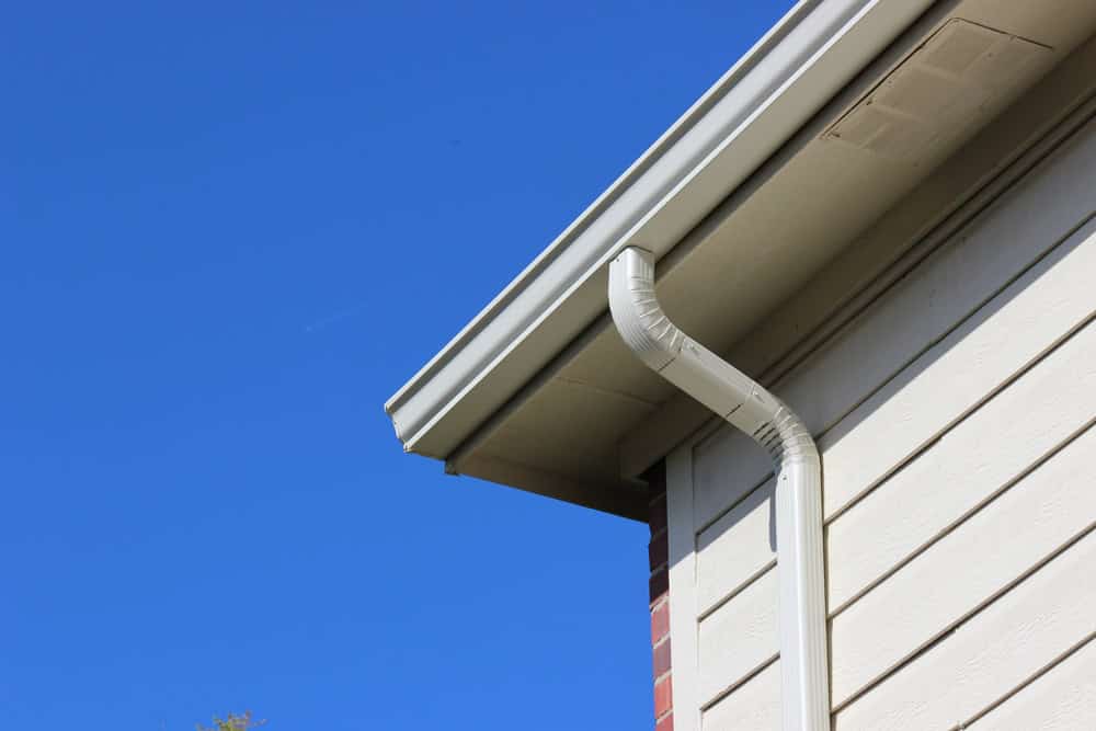 Close-up of a house corner in New London County, CT, showcasing a light-colored rain gutter system against a clear blue sky. The downspout seamlessly runs down the siding, reflecting expert roof installation craftsmanship.