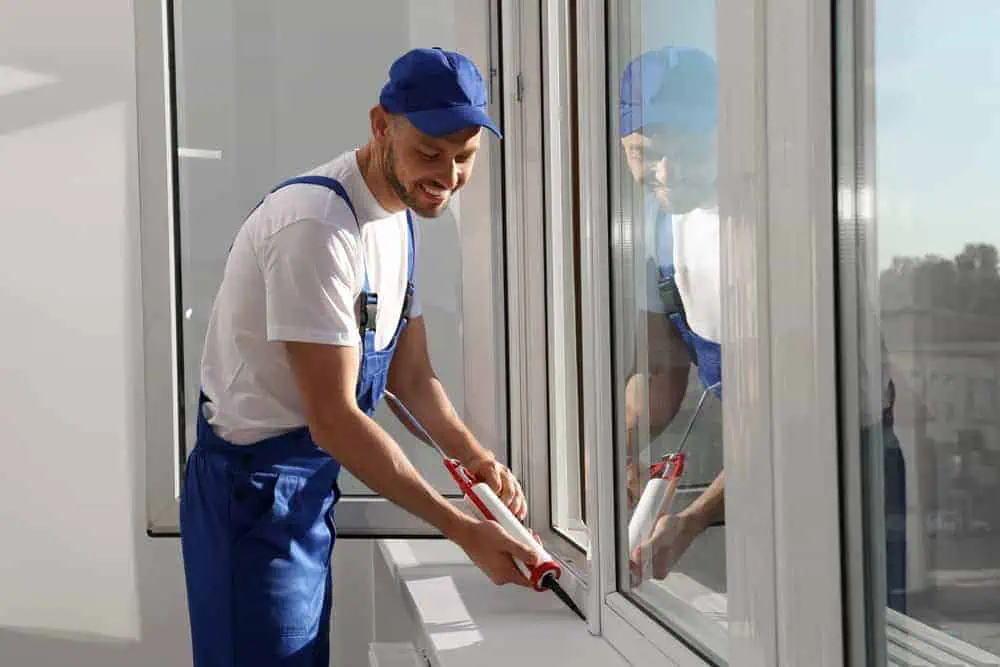 A man in blue overalls and a cap is smiling while using a caulking gun to seal a window frame in New London County, CT. He stands in a bright room with natural light streaming in, perhaps taking a break from roof installation tasks.