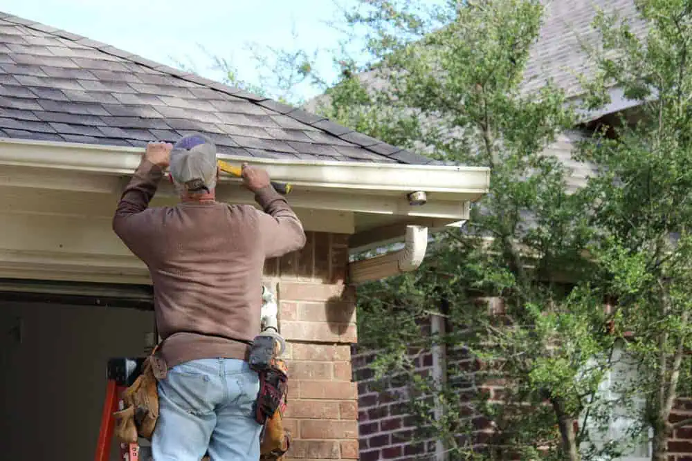 A roofing contractor in New London County stands on a ladder near a brick house, tool belt in place, carefully measuring the roof's edge with precision. Lush green trees serve as a serene backdrop to this scene of meticulous preparation for roof installation.