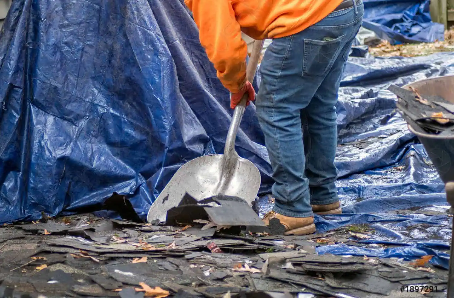 A person in an orange shirt and jeans, likely a roofing contractor in New London County, CT, uses a shovel to clear debris, including broken shingles, on a blue tarp. Fallen leaves are scattered among the debris.