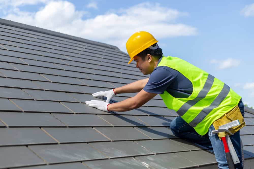 A roofing contractor in New London County, wearing a yellow hard hat and reflective vest, diligently repairs a roof with dark shingles. Against the backdrop of a clear blue sky, they skillfully use tools and wear white gloves for safety.
