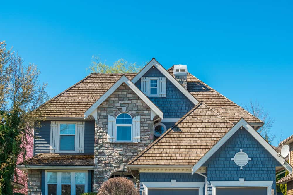 A charming two-story house in New London County, CT, boasts a blend of stone and shingle siding with arched and rectangular windows. Its gabled roof, expertly crafted by a local roofing contractor, is adorned with wooden shingles under a clear blue sky, framed by partially visible trees.