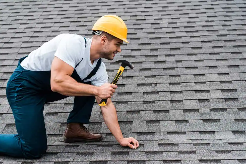 A person wearing a yellow hard hat and navy overalls is kneeling on a shingled roof in New London County, CT. Holding a hammer, they are focused and diligent as they work on the roof installation against a backdrop of clear blue sky.