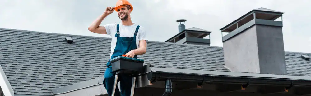 A construction worker, clad in a white shirt, blue overalls, and an orange hard hat, stands confidently on a roof near a chimney. As a skilled roofing contractor in New London County, he holds a toolbox while adjusting his hat under the cloudy sky and gray shingles.