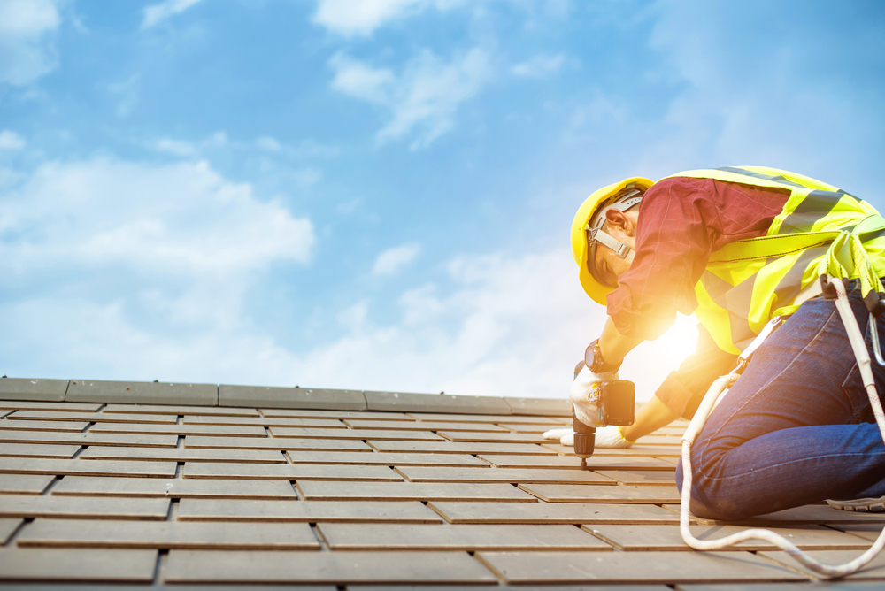 A construction worker wearing a yellow hard hat and reflective vest is kneeling on a roof, expertly handling a drill. Under the bright blue sky of New London County, CT, and with the sun shining in the background, he is secured with a safety harness while ensuring precise roof installation.