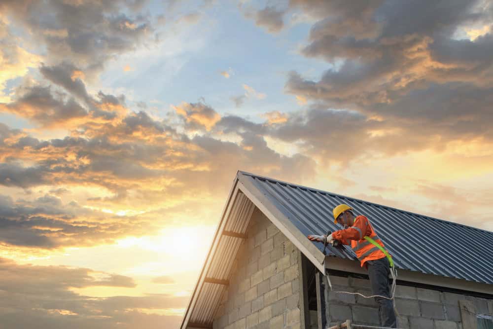 A roofing contractor in New London County, donned in a safety vest and helmet, diligently works on a rooftop at sunset. The partly cloudy sky glows with orange and yellow hues as the concrete blocks of the partially constructed building stand out against the horizon.
