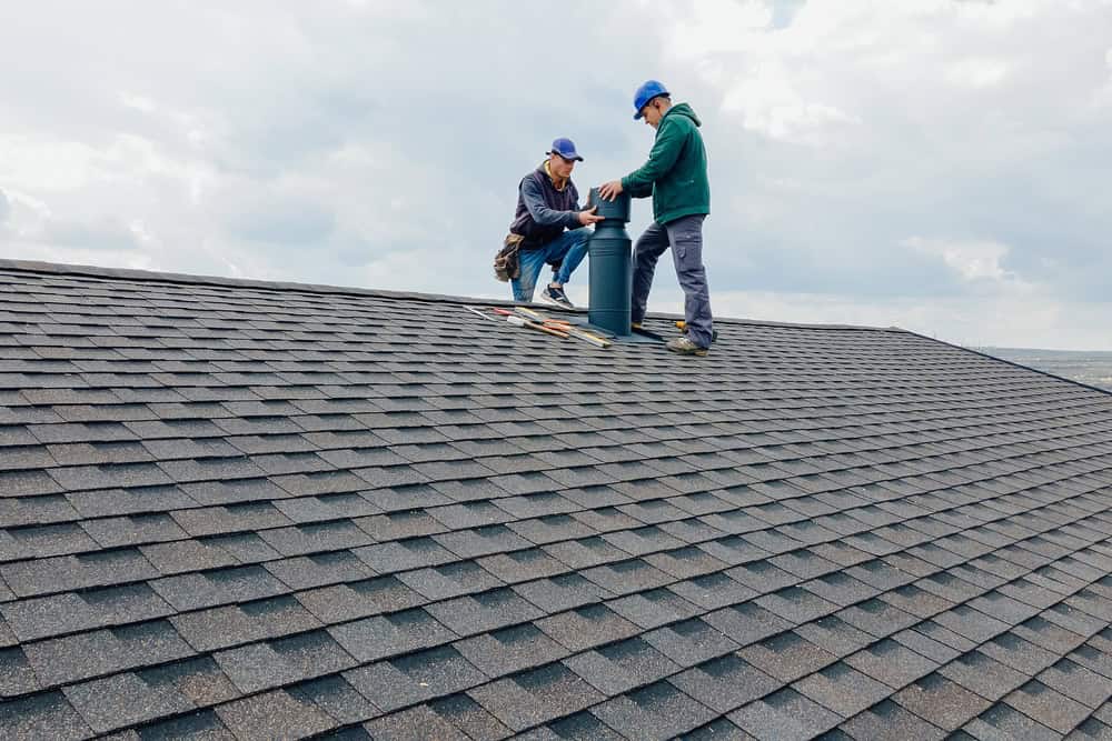 Two workers in blue caps and jackets skillfully execute a roof installation in New London County, securing a vent pipe on a shingled surface under a cloudy sky. One holds tools while the other adjusts the pipe, showcasing teamwork and precision on the high-pitched CT rooftop.