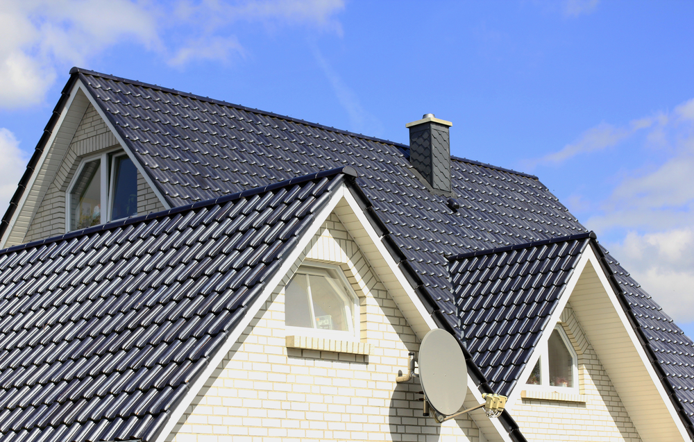 A house with white brick walls and a dark blue, tiled roof showcases the precision of roof installation in New London County, CT. It features three pointed gables with curved windows. A satellite dish is attached to one wall, and a chimney graces the roof under a clear blue sky with a few clouds.
