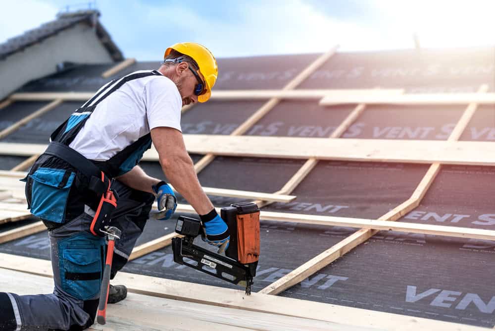 A construction worker in a yellow hard hat and protective gear expertly uses a nail gun to secure wooden beams on a roof. The scene of this roof installation in New London County, CT offers a bright, slightly cloudy sky as the perfect backdrop.