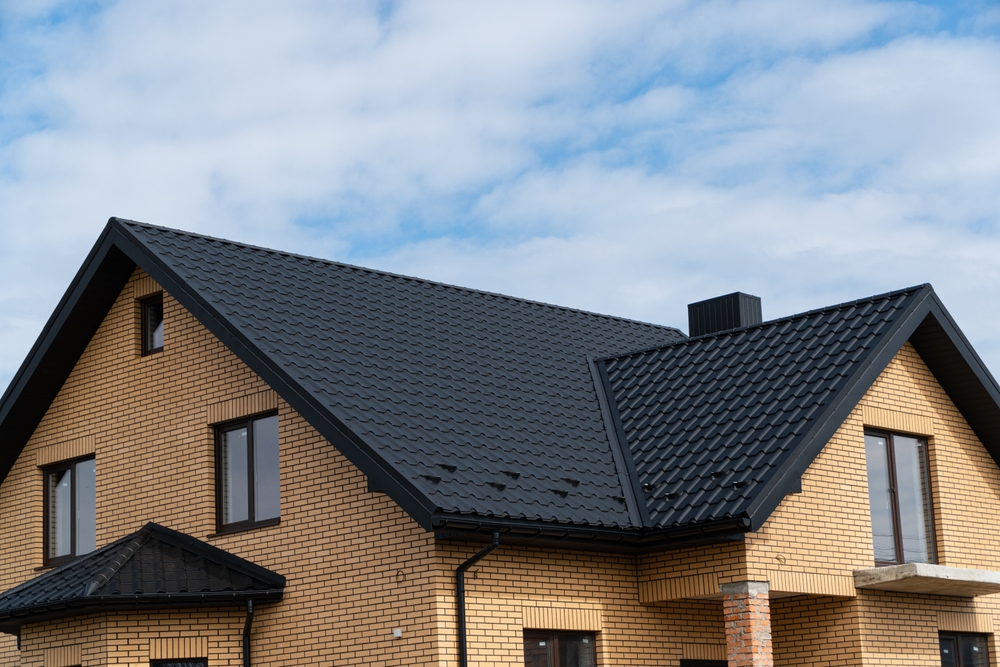 A modern brick house with a dark, pitched roof against a partly cloudy sky stands proudly in New London County. The home, serviced by a top-tier roofing contractor, showcases large windows, sleek black trim, and a prominent chimney.