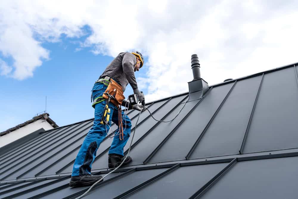 A roofing contractor in New London County, CT, is wearing safety gear as they carefully install or inspect a metal roof under a partly cloudy sky. Equipped with tools and harnesses, they remain focused on their task.