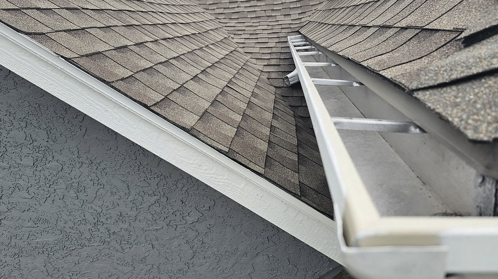 A close-up view showcases a New London County roof installation with brown asphalt shingles. The white gutter remains debris-free as a metal ladder rests at the edge, hinting at maintenance by a skilled roofing contractor in CT.