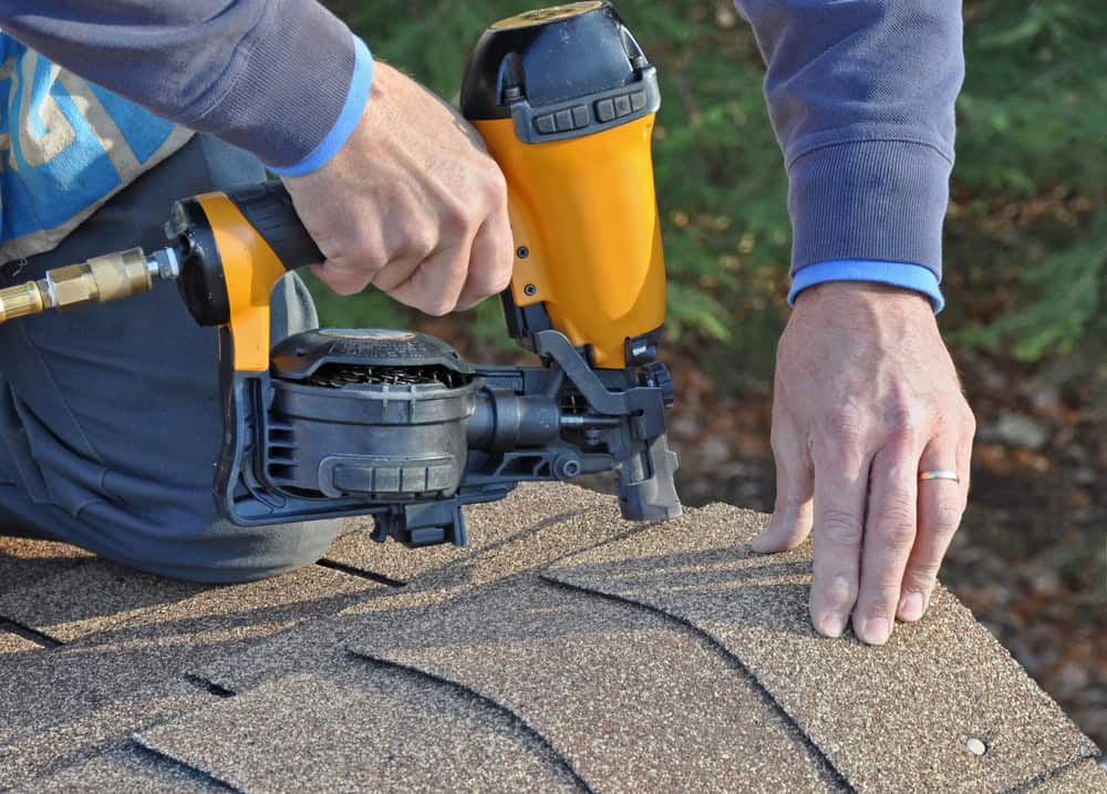A person adeptly uses a yellow nail gun to install asphalt shingles, demonstrating expert roof installation in New London County, CT. One hand deftly holds the tool while positioning the shingles with precision. In the background, a blurred glimpse of greenery adds a touch of nature.