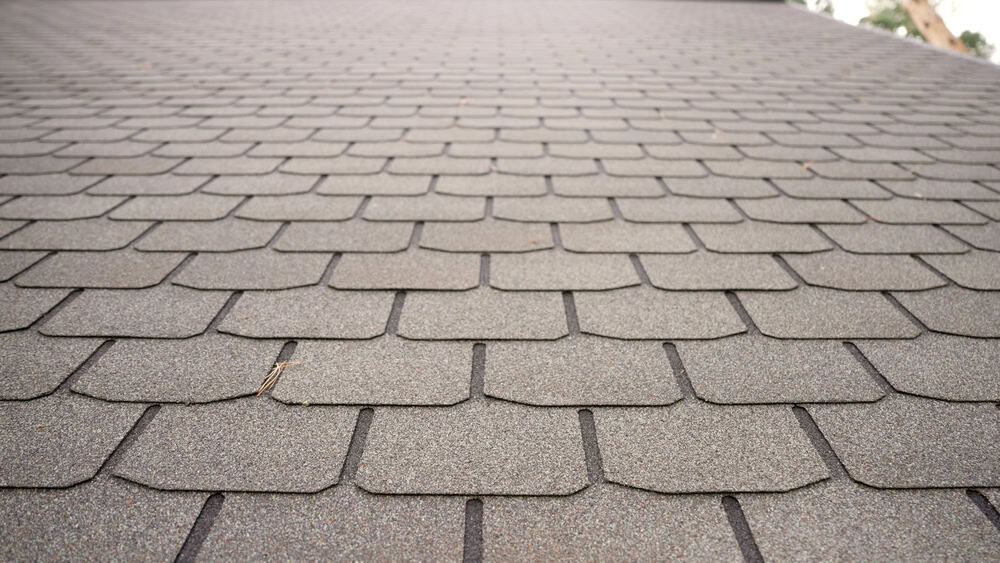 Close-up of a gray asphalt shingle roof in New London County, CT, reveals an overlapping pattern of rectangular shingles. The rough texture, dotted with tiny grains and scattered twigs, highlights the craftsmanship you’d expect from a skilled roofing contractor in CT.