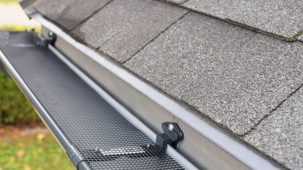 Close-up of a house gutter system on a gray shingled roof in New London County, CT. A black mesh guard is securely attached, effectively preventing debris from entering the gutter. A hint of greenery enhances the serene scene. Perfect for seamless roof installations in CT.