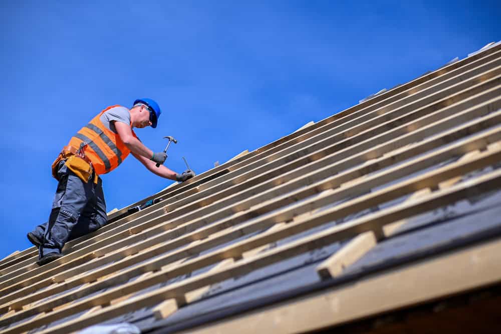 A roofing contractor in New London County, wearing an orange safety vest and blue helmet, is expertly hammering nails into the wooden framework of a sloped roof under a clear blue CT sky.