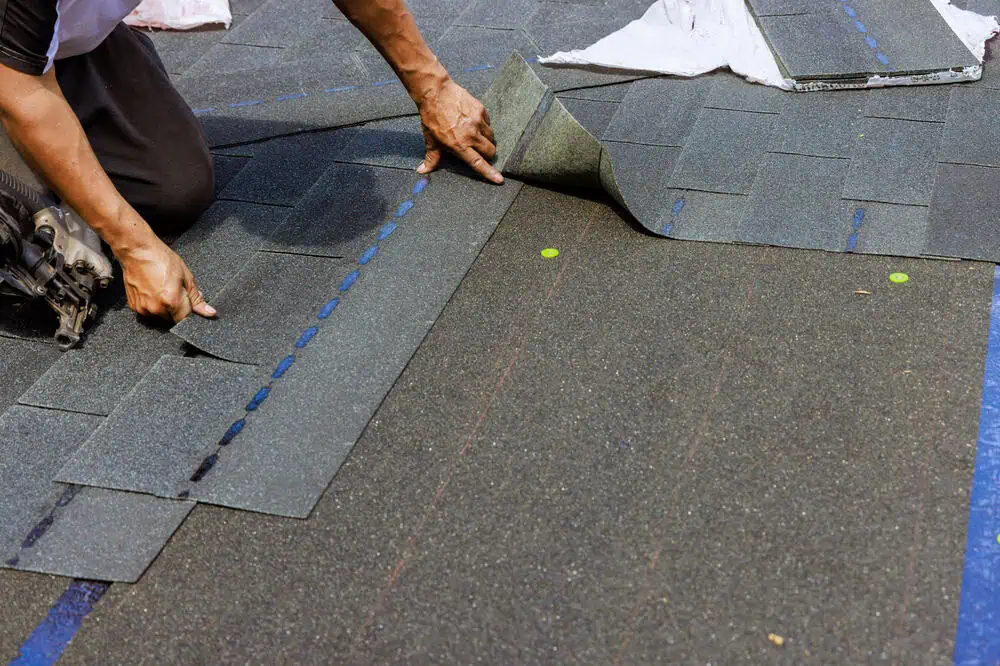 A roofing contractor in New London County, CT, is installing dark shingles on a roof with precision. Using a nail gun, they adjust each shingle for perfect alignment. The surface is marked with blue and red guidelines, complemented by small yellow indicators on the shingles.